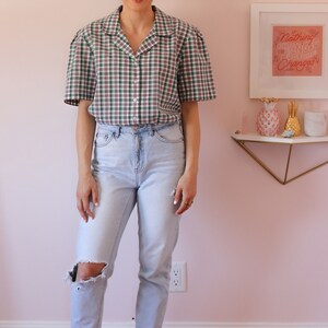 May include: A woman wearing a green and white plaid button-up shirt with a light blue pair of jeans and white sneakers. The shirt has a relaxed fit and the jeans have a distressed look. The woman is standing in front of a pink wall with a framed print that says "Nothing Changes, Nothing Changes".