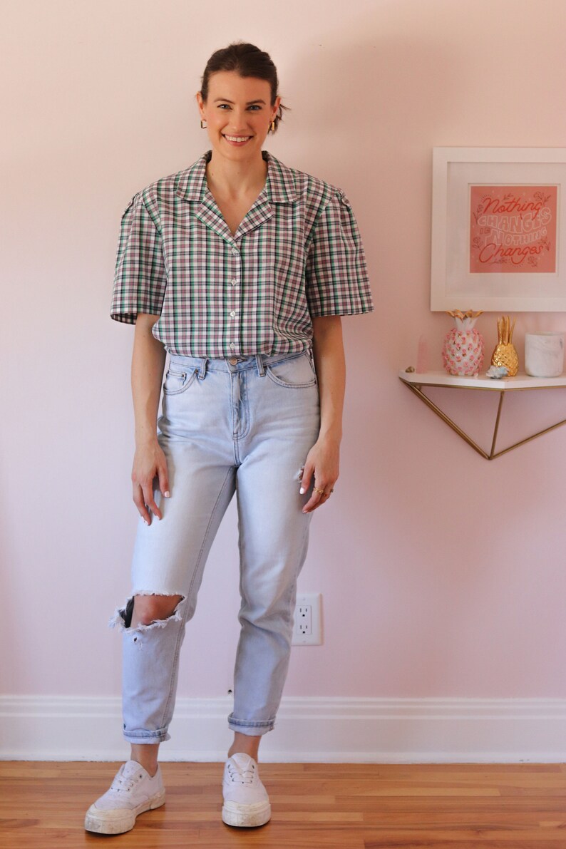 May include: A woman wearing a green and white plaid button-up shirt with a light blue pair of jeans and white sneakers. The shirt has a relaxed fit and the jeans have a distressed look. The woman is standing in front of a pink wall with a framed print that says "Nothing Changes, Nothing Changes".