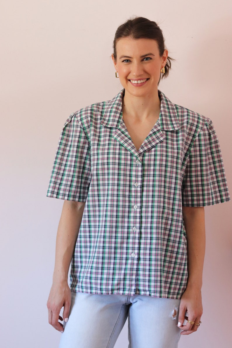 May include: A woman wearing a white, green, and black plaid button-down shirt with short sleeves. She is wearing light blue jeans.