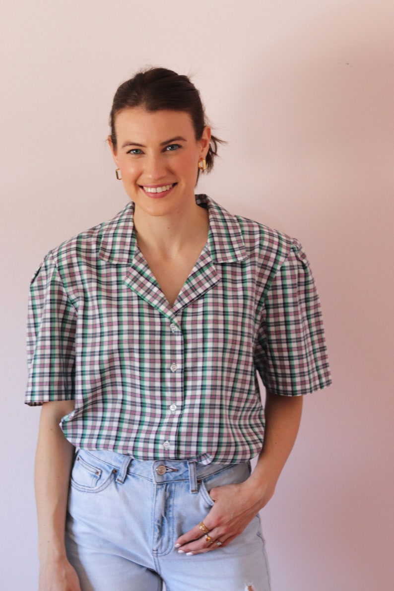 May include: A woman wearing a short-sleeved, green, white, and pink plaid button-down shirt with a light blue pair of jeans. She is smiling and has her hand in her pocket.