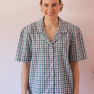 May include: A woman wearing a white, green, and pink plaid short-sleeved button-up shirt with a collar. She is wearing light blue jeans and has her hair pulled back.