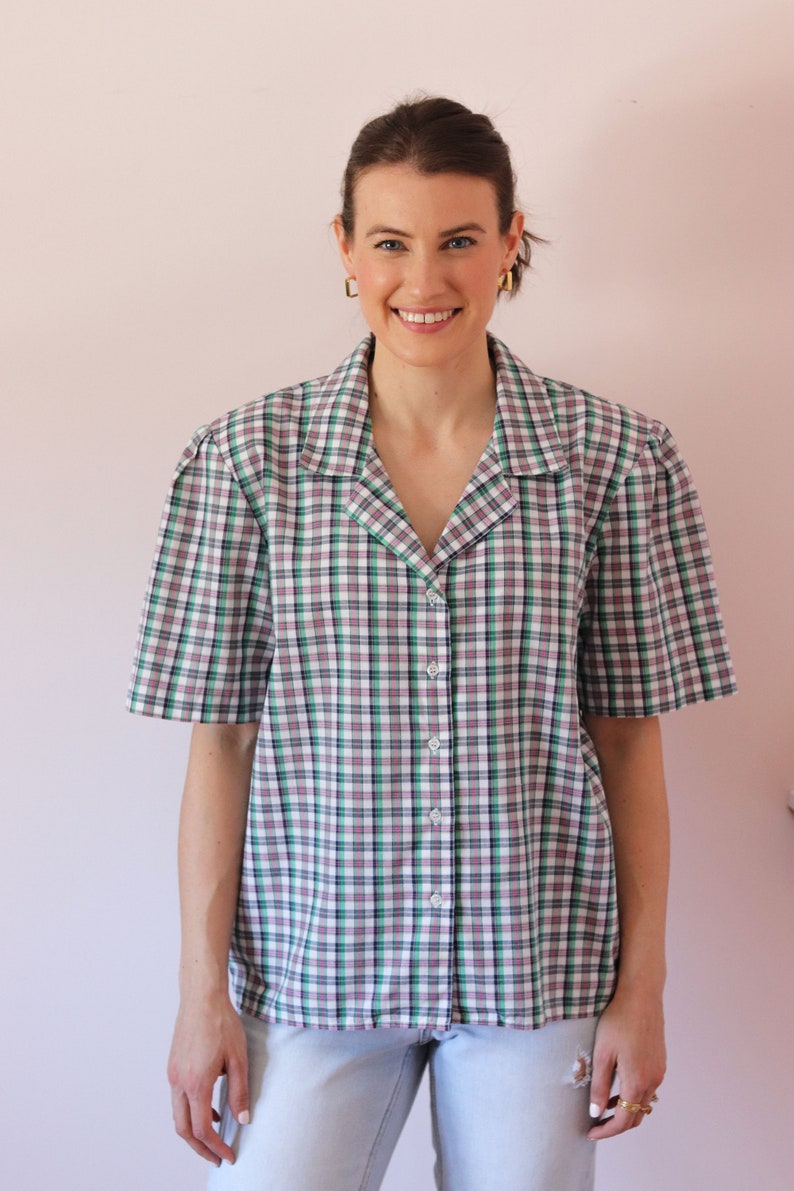 May include: A woman wearing a white, green, and pink plaid short-sleeved button-up shirt with a collar. She is wearing light blue jeans and has her hair pulled back.
