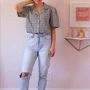 May include: A woman wearing a green, white, and purple plaid short-sleeved button-up shirt with a light blue pair of jeans that have a distressed knee. She is wearing white sneakers. The background is a light pink wall with a white frame on the wall with the text "Nothing Changes Until Something Changes".