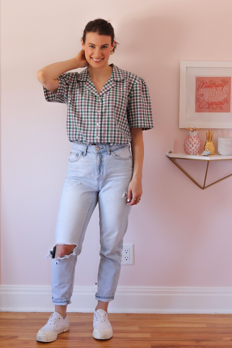 May include: A woman wearing a green, white, and purple plaid short-sleeved button-up shirt with a light blue pair of jeans that have a distressed knee. She is wearing white sneakers. The background is a light pink wall with a white frame on the wall with the text "Nothing Changes Until Something Changes".