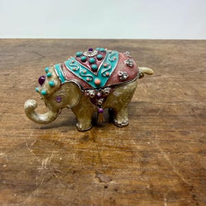 May include: A decorative elephant figurine with a detailed, colorful design. The elephant is tan with a pink and turquoise saddle adorned with purple and turquoise accents. The figurine has a small, curved trunk and a small tail.