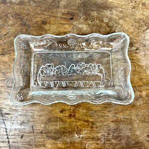 May include: A clear glass rectangular dish with a scalloped edge. The center of the dish features a relief of the Last Supper scene. The edges are decorated with grape vines and other floral elements. The dish is on a wooden surface.