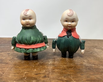 May include: Two antique bisque dolls with bald heads and painted faces. One doll wears a green dress with a coral-colored trim, the other wears a green outfit with a red jacket. Both dolls have small arms and legs and are standing on a wooden surface.
