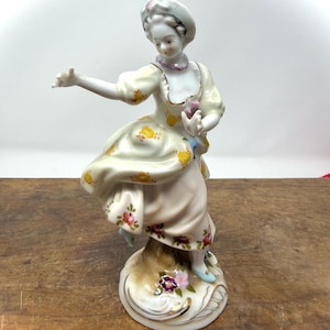 May include: A porcelain figurine of a woman in a flowing dress, holding a small object. The dress is cream-colored with yellow and pink floral accents. The figurine is on a decorative base with gold trim and additional floral details.