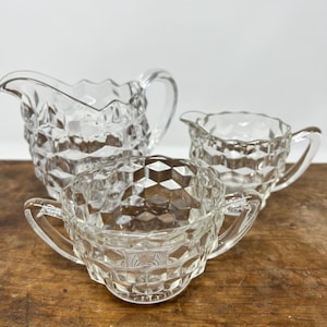 May include: A clear glass creamer and sugar bowl set. The creamer and sugar bowl have a diamond pattern. The sugar bowl has two handles. The set is on a wooden surface.
