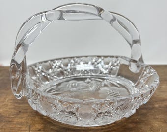 May include: Clear glass basket with a twisted handle. The basket features a floral design on the base and a diamond-cut pattern around the rim. The basket is sitting on a wooden surface.