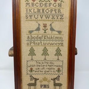 May include: Framed cross-stitch sampler with a dark wood frame. The sampler features an alphabet, birds, trees, deer, and the text "This is the day which the Lord hath made; we will rejoice and be glad in it."
