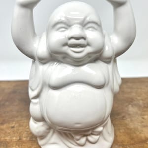 May include: A white ceramic Buddha figurine with arms raised. The smiling Buddha has a large belly and is wearing a robe. The figurine is a decorative item, likely for home decor or spiritual practice.