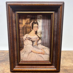 May include: A framed portrait of a woman in a white dress with a light blue bodice. The dark brown wooden frame surrounds the image, which is set against a backdrop of a wooden door and a curtain.