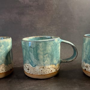 May include: Three handmade ceramic mugs. The mugs feature a turquoise glaze on the upper section, blending into a speckled white and brown base. Each mug has a rounded handle and a rustic, artisanal appearance.