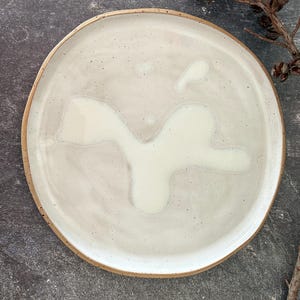 May include: A round ceramic plate with a speckled, off-white glaze and a natural brown rim. The plate has a unique, organic shape and a smooth surface. The plate is suitable for serving food.