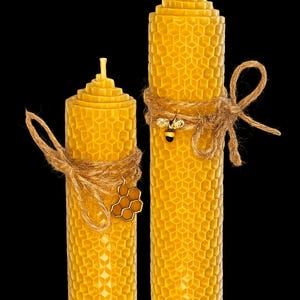 May include: Two yellow beeswax candles with a honeycomb pattern. The candles are adorned with twine and bee-themed charms. The candles are of different heights, with a black background.