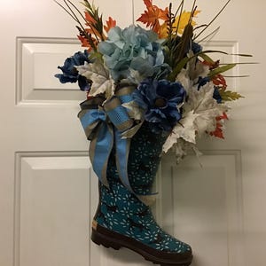 May include: A blue rain boot with a floral pattern is filled with a bouquet of blue, white, and orange flowers. The boot is decorated with a blue and gold ribbon.