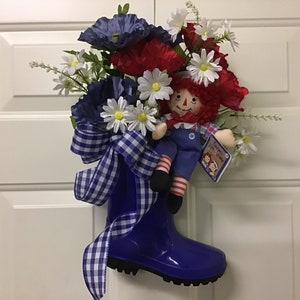 May include: A blue rubber rain boot with a red, white, and blue floral arrangement and a rag doll sitting inside. The boot has a blue and white checkered ribbon tied around it.