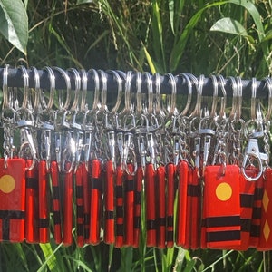 May include: A collection of red keychains with silver rings and clasps. Each keychain has a rectangular shape with a yellow circle and black stripes. The keychains are displayed on a black metal rack against a backdrop of green foliage.