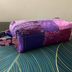 Peut inclure: Une pochette zippée en tissu patchwork violet avec une anse en tissu floral. La pochette est posée sur une surface à motifs verts et dorés.
