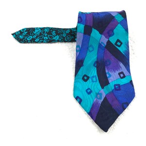 Puede incluir: Una corbata de seda con estampado geométrico azul, morado y negro. La corbata tiene una punta con estampado floral.
