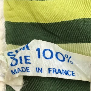Może przedstawiać: Biała metka z tkaniny z niebieskim tekstem, kt&oacute;ry brzmi "Silk Die 100% Made in France". Metka jest przymocowana do zielono-ż&oacute;łtej tkaniny w paski.