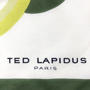 Może przedstawiać: Biały jedwabny szal z abstrakcyjnym wzorem w kolorze zielonym i ż&oacute;łtym. Na szalu nadrukowany jest napis "TED LAPIDUS PARIS".