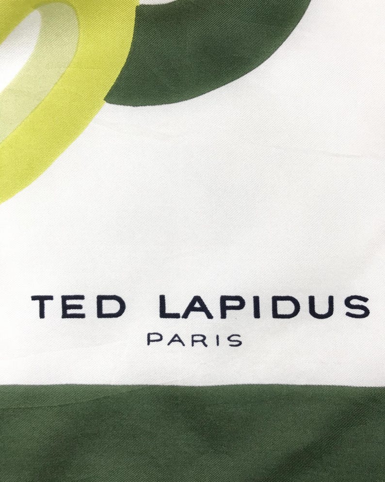 Może przedstawiać: Biały jedwabny szal z abstrakcyjnym wzorem w kolorze zielonym i ż&oacute;łtym. Na szalu nadrukowany jest napis "TED LAPIDUS PARIS".