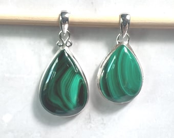 May include: Two teardrop-shaped malachite pendants with silver-colored settings. The pendants are a vibrant green with dark green swirling patterns. They hang from a wooden dowel against a white background.