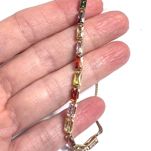 May include: A gold necklace with a series of rectangular, multi-coloured gemstones. The gemstones are arranged in a linear pattern, displaying a spectrum of colours including green, purple, yellow, and red. The necklace is set against a white background.