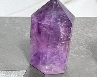 May include: A faceted amethyst crystal point, showcasing a gradient of purple hues. The crystal is cut into a hexagonal shape, with a flat base and a pointed top. The polished surface reflects light, highlighting the natural variations in color.