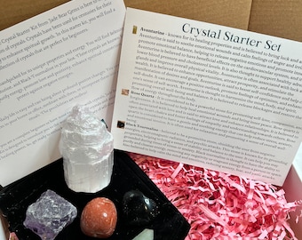 May include: A crystal starter kit with a black velvet tray containing seven crystals: a clear selenite tower, a green aventurine, a purple amethyst, a red rose quartz, a black tourmaline, a clear selenite stick, and a green aventurine stick. The kit is packaged in a pink and white shredded paper box. The box has a label with the text "Crystal Starter Set" and descriptions of the crystals.