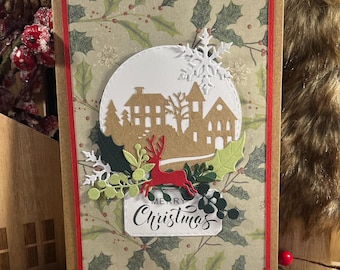 Könnte beinhalten: Handgemachte Weihnachtskarte mit festlichem Design. Die Karte zeigt einen kreisförmigen Ausschnitt einer Dorflandschaft, ein rotes Rentier und die Worte "Merry Christmas". Die Karte ist von dekorativen Elementen umgeben, darunter Schneeflocken und Stechpalmen.