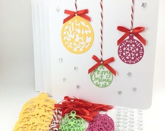 May include: A set of Christmas card making supplies with white cards, red and white twine, red ribbon, yellow, green, and pink paper ornaments, silver glitter stars, and silver beads.