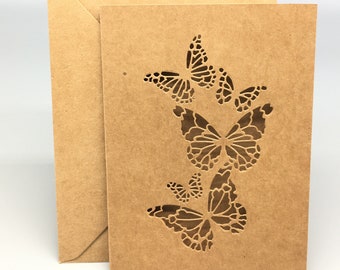 Puede incluir: Una tarjeta marrón con un sobre blanco. La tarjeta tiene un diseño recortado de tres mariposas.
