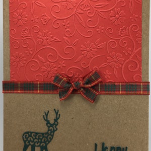 Puede incluir: Una tarjeta marr&oacute;n con papel estampado rojo en la parte superior. Una cinta a cuadros con un lazo est&aacute; atada alrededor de la tarjeta. Una silueta verde de un reno y el texto "Feliz Navidad" est&aacute;n en la parte inferior de la tarjeta.