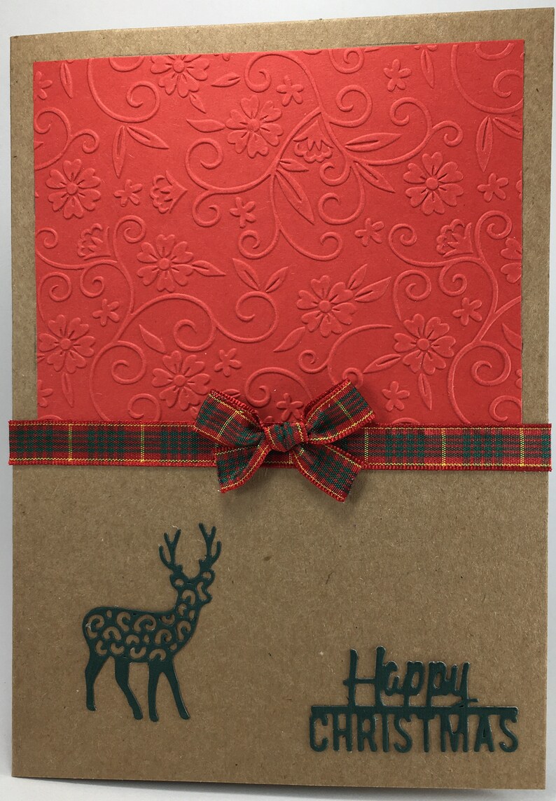 Puede incluir: Una tarjeta marr&oacute;n con papel estampado rojo en la parte superior. Una cinta a cuadros con un lazo est&aacute; atada alrededor de la tarjeta. Una silueta verde de un reno y el texto "Feliz Navidad" est&aacute;n en la parte inferior de la tarjeta.