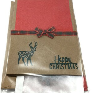 Puede incluir: Una tarjeta de Navidad con una cubierta de papel marr&oacute;n, una tarjeta roja en el interior y una cinta a cuadros. La parte delantera de la tarjeta presenta una silueta negra de un reno con un estampado de leopardo y el texto "Feliz Navidad".
