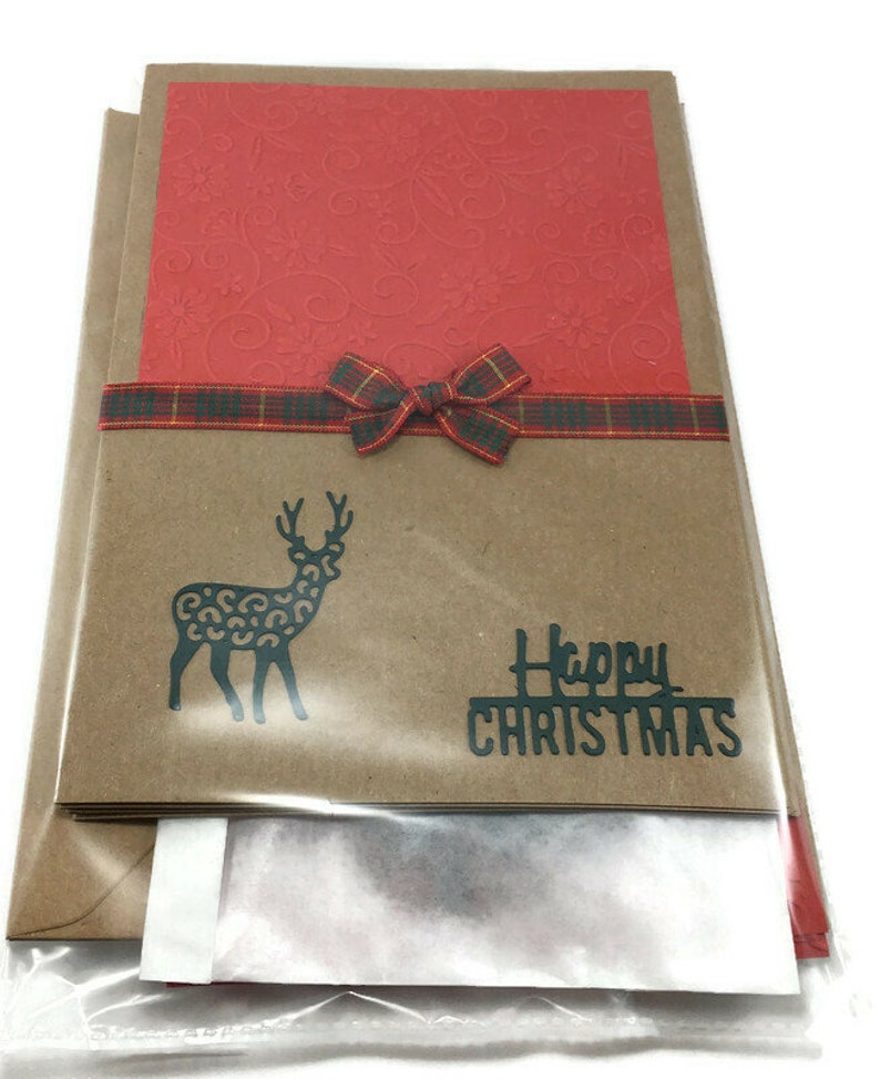 Puede incluir: Una tarjeta de Navidad con una cubierta de papel marr&oacute;n, una tarjeta roja en el interior y una cinta a cuadros. La parte delantera de la tarjeta presenta una silueta negra de un reno con un estampado de leopardo y el texto "Feliz Navidad".