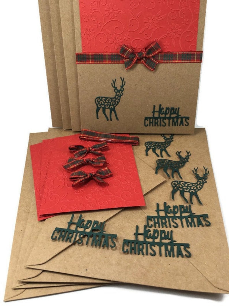 Puede incluir: Un conjunto de tarjetas de Navidad de papel kraft marr&oacute;n con sobres rojos. Las tarjetas presentan una silueta negra de un reno y el texto "Feliz Navidad". Los sobres tienen un patr&oacute;n en relieve rojo y est&aacute;n decorados con lazos de cinta de tart&aacute;n verde y rojo.