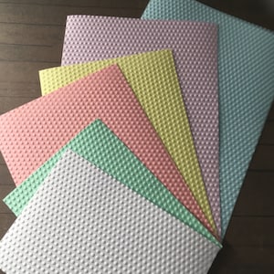 Op de afbeelding: Een verzameling van getextureerde papieren vellen in verschillende pastelkleuren, waaronder roze, geel, groen en blauw. Elk vel heeft een herhalend stippenpatroon, waardoor een tastbaar oppervlak ontstaat. De papieren zijn in een waaier gerangschikt, waarbij de verschillende tinten worden getoond.