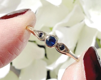 May include: A gold ring featuring three dark blue gemstones. The central stone is round, flanked by two smaller, oval-shaped stones. The ring's band is thin and delicate, with intricate detailing around the gemstones.