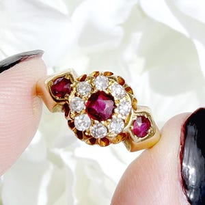 May include: A gold ring featuring a central square-cut red ruby surrounded by a halo of small round white diamonds. The band includes two additional red rubies, one on each side of the central cluster, set within a polished yellow gold metal frame.