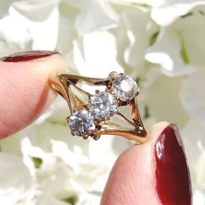 May include: A gold ring with three clear gemstones. The ring has a unique geometric design with a diamond-shaped center holding the gemstones. The gemstones are round and set in a gold band. The ring is held up against a background of white flowers.