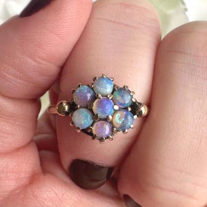 May include: A cluster ring featuring multiple opals in a gold setting. The opals display a range of colors, including blue, purple, and white. The ring's design is intricate, with small details around each opal. The ring is held by a hand.