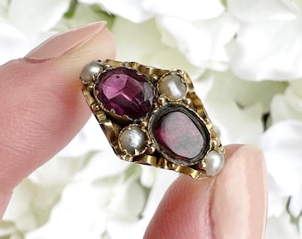 May include: A vintage gold-toned brooch with a diamond shape, featuring two oval, deep red gemstones and several small, round pearls. The brooch is held between fingers against a backdrop of white flowers.