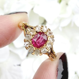 May include: A gold ring featuring a central oval ruby surrounded by a halo of smaller, clear gemstones. The ring's setting has an ornate, openwork design. The ring is held between two fingers against a blurred white floral background.