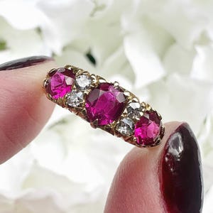 Puede incluir: Un anillo de oro con tres piedras de color rosa vibrante en corte ovalado que alternan con dos piedras transparentes más pequeñas en corte redondo. Las piedras están engastadas en una fila horizontal en una banda de oro amarillo pulido.