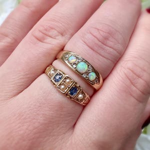 Op de afbeelding: Twee gouden ringen. De bovenste ring heeft drie opalen en kleine diamanten. De onderste ring heeft vierkante blauwe stenen en kleine parels. Beide ringen zitten aan een vinger.