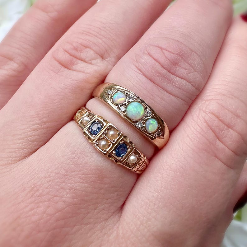 Op de afbeelding: Twee gouden ringen. De bovenste ring heeft drie opalen en kleine diamanten. De onderste ring heeft vierkante blauwe stenen en kleine parels. Beide ringen zitten aan een vinger.